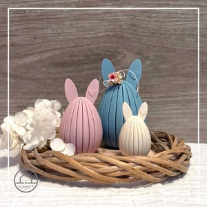 Può includere: Tre decorazioni a forma di uovo di Pasqua a forma di coniglietto in un cestino di vimini. Le uova sono rosa, blu e color crema, ognuna con orecchie da coniglio. Fiori bianchi a sinistra. Lo sfondo è una superficie di legno.