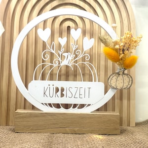 Puede incluir: Decoración metálica blanca con temática de calabaza con el texto alemán "Kürbiszeit" sobre una base de madera. El diseño incluye una calabaza, corazones y una pequeña calabaza de madera con flores secas. El fondo es un arco de madera.