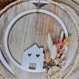 May include: A white house-shaped decoration with heart cutouts and a wooden tag reading "Lieblings Platz". The house is within a white circle and a wooden heart-shaped frame. The design is completed with dried flowers and wooden beads, creating a rustic aesthetic.