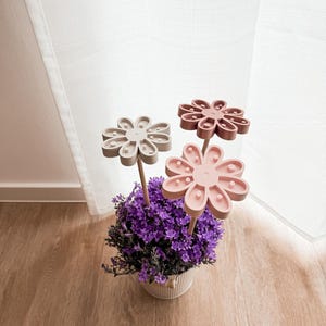 Può includere: Composizione floreale decorativa con tre ornamenti a forma di fiore nei toni del rosa e del grigio su steli di legno. I fiori sono in un vaso bianco con fiori viola, davanti a una tenda bianca e un pavimento in legno.