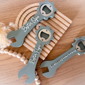 Può includere: Tre apribottiglie grigi a forma di chiave inglese con apribottiglie argentati. Due apribottiglie hanno il testo "Bester Opa" e "Bester Papa" in scrittura bianca. Il terzo apribottiglie ha il testo "Lieblingswerkzeug".