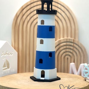 Peut inclure: Un phare décoratif avec des rayures bleues et blanches, un sommet noir et une base noire. Le phare est exposé sur une surface en bois, avec un objet en forme de maison blanche et des arches en bois en arrière-plan.
