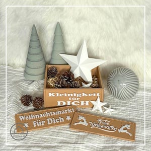 Może przedstawiać: Zestaw dekoracji świątecznych, w tym dwa jasnozielone drzewka w kształcie stożka, biała gwiazda i drewniane pudełko wypełnione szyszkami. Dwa znaki głoszą "Weihnachtsmarkt für Dich" i "Frohe Weihnachten". Obecna jest również szara, żebrowana kula.
