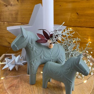 May include: Two sage green horse figurines with embossed floral patterns, one featuring a central hole holding a tall white candle and dried foliage. A silver star ornament and white baby's breath flowers are arranged nearby.