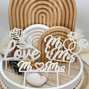 Könnte beinhalten: Weiße, lasergeschnittene Tortenaufsätze mit den Worten "love" und "Mr & Mrs" sowie einem Diamantring-Design. Die Aufsätze werden auf einem weißen Tablett mit Holzperlen präsentiert, mit einem hölzernen Regenbogenbogen im Hintergrund. Hochzeitsthema.