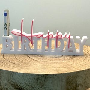 Könnte beinhalten: Ein weißer „Happy Birthday“-Kuchenaufsatz mit rosa Akzenten. Das Wort „Happy“ ist kursiv geschrieben, und „Birthday“ in Großbuchstaben. Eine aufgerollte Banknote befindet sich im Halter. Der Aufsatz steht auf einer Holzoberfläche.
