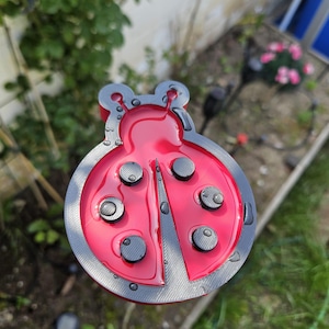 May include: A red and gray ladybug-shaped garden stake. The ladybug has a glossy red body with black circular spots and a gray textured border. Water droplets are visible on the surface, suggesting recent watering or rain. The stake is designed for outdoor garden decor.