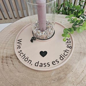 May include: A wooden candle holder with the text "Wie schön, dass es dich gibt" and a black heart. The holder has a glass cylinder with a pink candle and white stones.