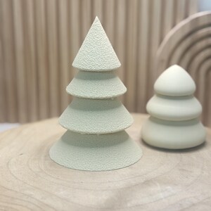May include: Two light green decorative Christmas trees. One is a layered cone, the other a rounded shape. The trees are on a wooden surface with a light brown background.