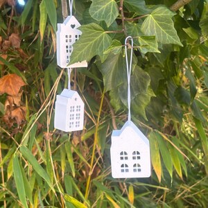 May include: Three small, white, house-shaped ornaments suspended from white strings against green foliage. Each ornament has cut-out windows and a heart-shaped detail. The ornaments are hanging from a tree branch.