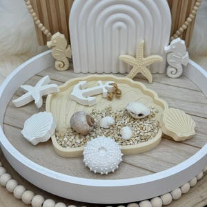 Puede incluir: Una bandeja redonda blanca con temática playera. Presenta un plato en forma de concha lleno de arena, conchas y un ancla blanca. Caballitos de mar decorativos, estrellas de mar y un arco iris crean una estética costera.