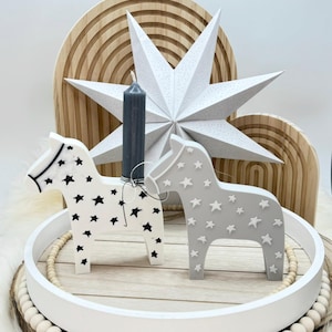 May include: A decorative arrangement featuring two wooden horse figurines, one white with black stars and the other gray with white stars. A gray candle sits between them, with a white star and wooden arch in the background.
