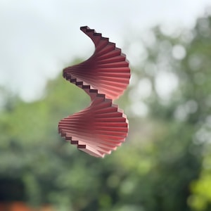 Puede incluir: Un móvil eólico de madera en forma de espiral, en tonos rosa y marrón. El diseño presenta segmentos superpuestos en forma de abanico que crean una forma retorcida y simétrica. El móvil está suspendido sobre un fondo verde borroso. Medidas en centímetros.