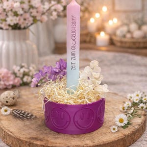 Könnte beinhalten: Eine brennende rosa-blaue Kerze mit dem Text "ZEIT ZUM GLÜCKLICHSEIN" steht in einem lila Behälter mit Smiley-Gesichtern. Der Behälter enthält Trockenblumen und steht auf einer Holzscheibe, mit Blumen im Hintergrund.