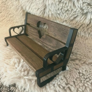 Puede incluir: Un pequeño banco de madera con detalles en metal negro. El banco tiene un recorte en forma de corazón en la parte posterior y detalles en forma de corazón en los lados. El banco está hecho de madera oscura y tiene un aspecto rústico.