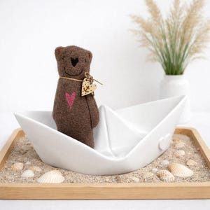 Può includere: Un orsacchiotto di feltro marrone con un cuore rosa sul petto, in una barca di carta bianca. L'orso ha un'etichetta di legno a forma di cuore con un'ancora. La barca è in un vassoio di legno con sabbia e conchiglie.