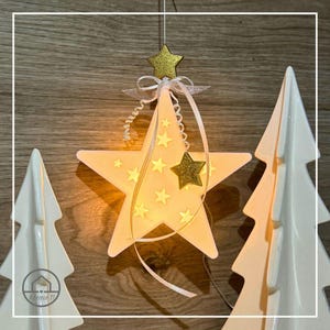 May include: A glowing star-shaped Christmas ornament with smaller star cutouts and a gold glitter star accent. The star is suspended by a thin string and adorned with a white ribbon. Two white ceramic Christmas trees flank the star.