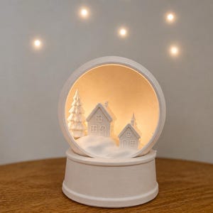 May include: A circular white decorative light featuring a miniature winter scene inside. The scene includes two small white houses and a stylised white pine tree resting on a snow-covered base. The interior is illuminated with a warm glow.