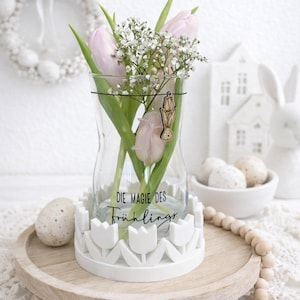 May include: A clear glass vase with pink tulips and white baby's breath, set on a white tulip-shaped base. The vase has the text "DIE MAGIE DES Frühlings". The arrangement sits on a wooden tray with decorative Easter eggs.