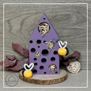 Può includere: Una scatola decorativa a forma di casa in legno viola con ritagli circolari e un tetto triangolare. La scatola è adornata con ornamenti a forma di ape gialla e nera con ali a forma di cuore bianco. La scatola poggia su una fetta di legno.