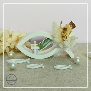 Könnte beinhalten: Eine weiße, fischförmige Holzdekoration mit einem Kreuz und einer kleinen Glasflasche mit grünem Papier. Die Flasche ist mit Trockenblumen und einem Band verziert. Mehrere kleine weiße Fische und perlenartige Perlen sind um die Dekoration verstreut.