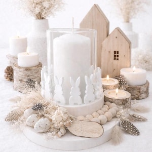 Op de afbeelding: Een witte kaarsenopstelling met een grote pilaar kaars in een glazen cilinder, omringd door witte konijnenfiguren. De scène bevat brandende kaarsen op houten schijven, miniatuur houten huisjes en decoratieve veren, wat een zachte, neutrale esthetiek creëert.