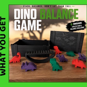 May include: A Dino Balance Game set featuring a black crate, a black curved balance base, and eight colourful dinosaur figures in red, green, and purple. The packaging text reads: STACK. BALANCE. DON'T LET THEM FALL. DINO BALANCE GAME. 8 DINOSAURS.