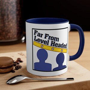 Puede incluir: Taza de café de cerámica blanca con borde y asa azules. La taza presenta un gráfico de dos siluetas azules con una herramienta de nivel amarilla sobre ellas. El texto "Far From Level Headed" está impreso en negro en la taza.