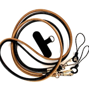 May include: A leather phone lanyard in black and tan. The lanyard has a black phone holder, gold-colored clasps, and black and white patterned loops. The leather cord is looped and coiled against a white background.