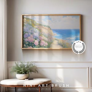 Op de afbeelding: Een ingelijste kunstdruk toont een kustlandschap met een pad naar de zee. Het kunstwerk toont een heuvel bedekt met roze en paarse bloemen, met een helderblauwe zee en lucht. De lijst is goudkleurig. De tekst "NINI ART BRUSH" staat onderaan.
