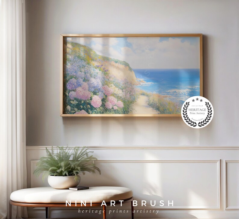 Op de afbeelding: Een ingelijste kunstdruk toont een kustlandschap met een pad naar de zee. Het kunstwerk toont een heuvel bedekt met roze en paarse bloemen, met een helderblauwe zee en lucht. De lijst is goudkleurig. De tekst "NINI ART BRUSH" staat onderaan.