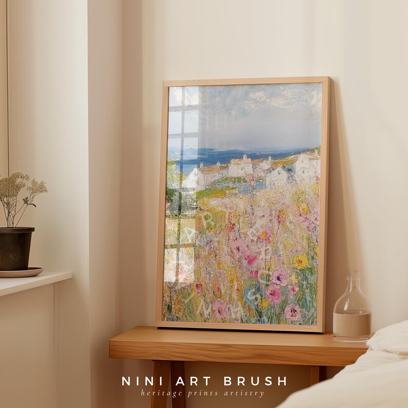 Op de afbeelding: Een ingelijst schilderij van een kustsc&egrave;ne met witte huizen en een veld met roze en gele bloemen. De lucht is blauw met witte wolken. Het schilderij is in een lichte houten lijst.