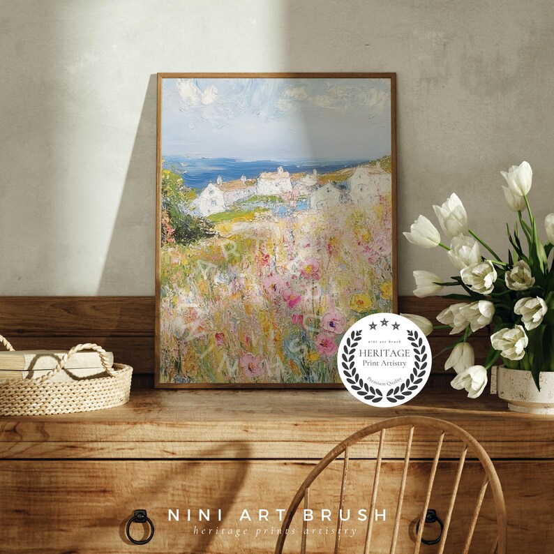Op de afbeelding: Een ingelijst schilderij van een kustsc&egrave;ne met witte huizen en een veld vol kleurrijke bloemen. Het schilderij is in een licht houten lijst en hangt aan een muur. Het schilderij is getiteld "NINI ART BRUSH" en heeft een "HERITAGE Print Artistry" zegel.