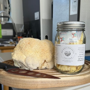 May include: A glass jar filled with dried lion's mane mushrooms. The jar has a white label with the text "Baja North Mercantile" and "Lion's Mane". The label also includes the text "25 Year Shelf Life" and "Net Wt 32 Grams Dry".