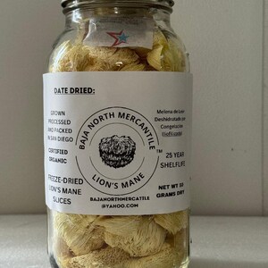 May include: A glass jar filled with freeze-dried lion's mane mushroom slices. The jar has a white label with the text "Baja North Mercantile" and "Lion's Mane". The label also includes information about the product, such as "Grown, Processed and Packed in San Diego", "Certified Organic", "Freeze-Dried Lion's Mane Slices", "25 Year Shelf Life", and "Net Wt 50 Grams Dry".
