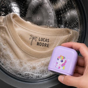Op de afbeelding: Een beige t-shirt met de zwarte tekst LUCAS MOORE en een palmboomicoon, geplaatst in een wasmachine. Een lichtpaarse vierkante stempelhouder met een kleurrijke eenhoorngrafiek wordt op de voorgrond van de afbeelding vastgehouden.