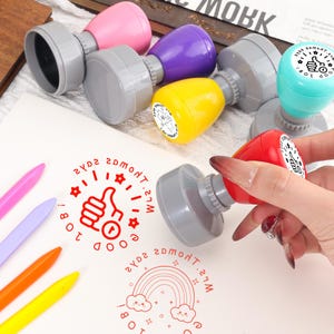 May include: A collection of colorful, plastic stamps with gray bases. The stamps are in various colors, including pink, purple, yellow, teal, and red. The stamped images include a thumbs-up, a rainbow, and the text "Good Job!"