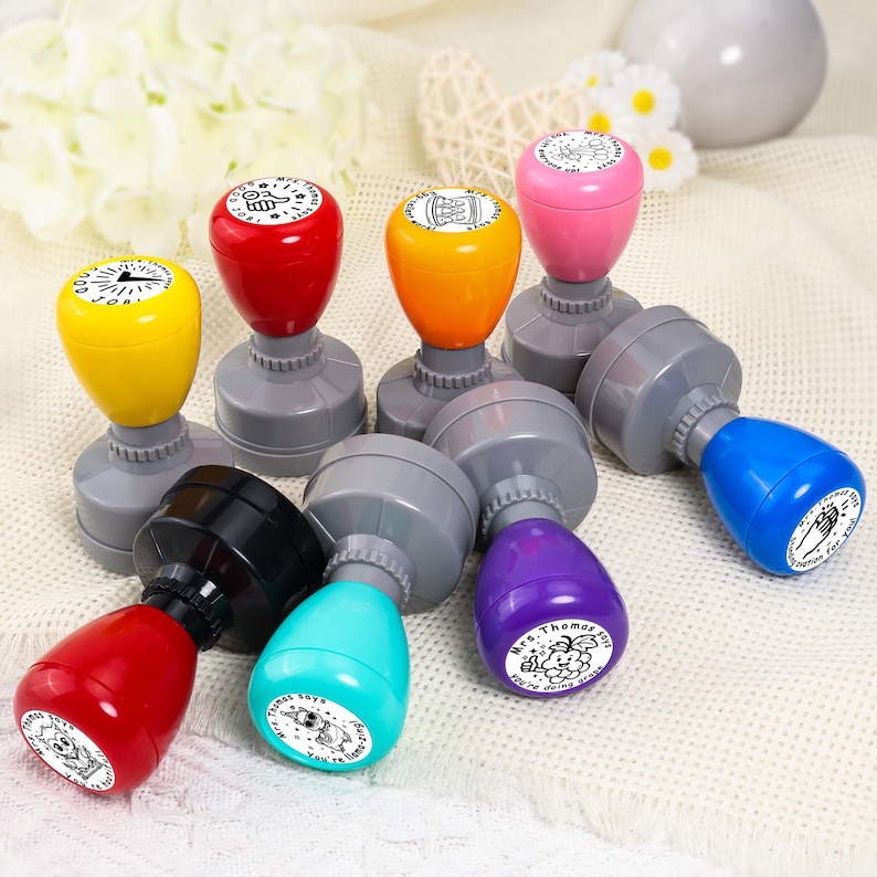 May include: A collection of colorful rubber stamps with gray bases. The stamps have various colors, including yellow, red, orange, pink, blue, black, teal, and purple. Each stamp has a unique design on its circular imprint, with text and illustrations.