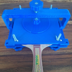 以下が含まれることがあります： 青い3Dプリントされた卓球ラケットプレスにねじクランプと、ハンドルに「Joola Cobra」の文字が書かれた木製卓球ラケットが付いている。