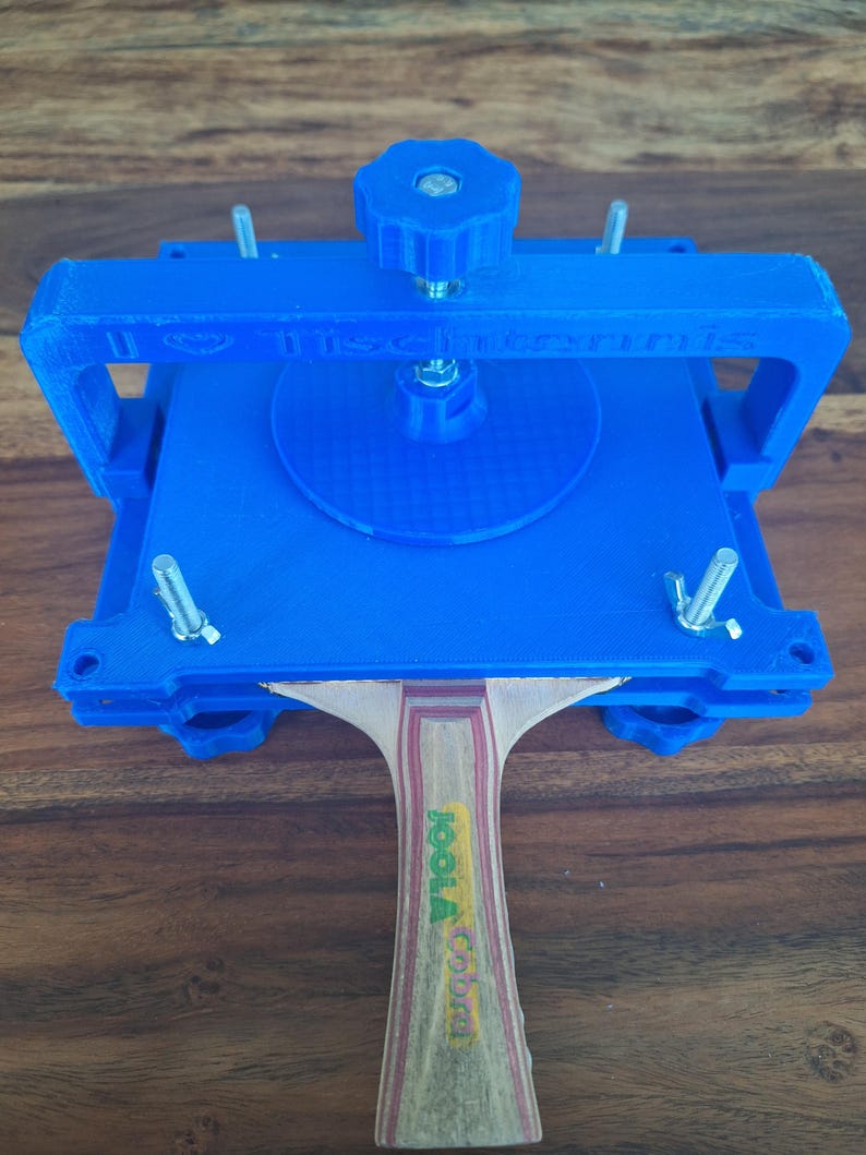 以下が含まれることがあります： 青い3Dプリントされた卓球ラケットプレスにねじクランプと、ハンドルに「Joola Cobra」の文字が書かれた木製卓球ラケットが付いている。