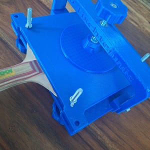 以下が含まれることがあります： 青い3Dプリントされた卓球ラケットプレスで、木製のラケットが挟まれています。プレスの上部にノブ、側面にネジがあります。プレスの側面には「TISCHTECHNIK」という文字が印刷されています。