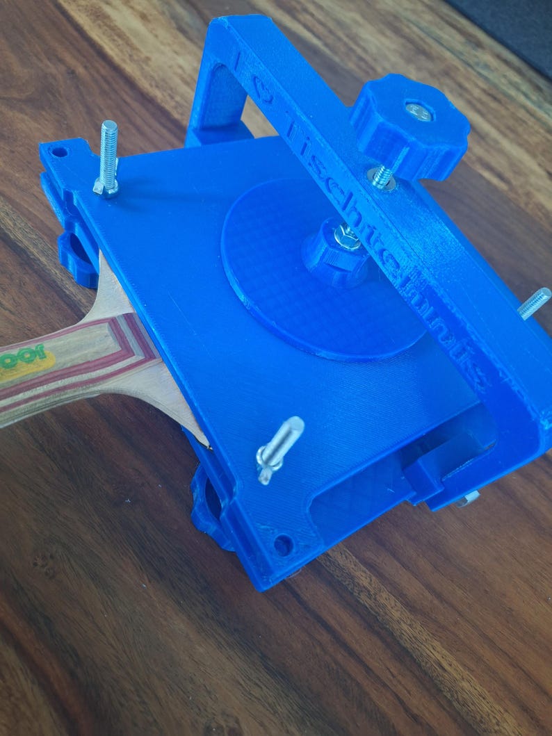 以下が含まれることがあります： 青い3Dプリントされた卓球ラケットプレスで、木製のラケットが挟まれています。プレスの上部にノブ、側面にネジがあります。プレスの側面には「TISCHTECHNIK」という文字が印刷されています。