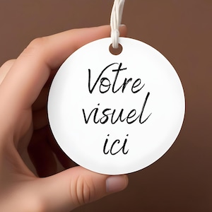 Op de afbeelding: Een witte, ronde tag met de Franse tekst "Votre visuel ici" in zwart schrift. De tag wordt door een hand vastgehouden tegen een bruine achtergrond, met een wit touwtje.