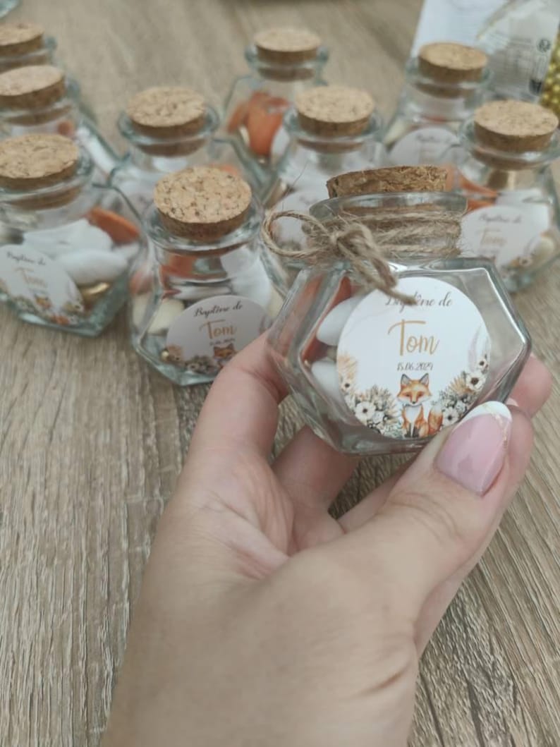 May include: Small glass jars with cork lids filled with white and orange candies. The jars have a fox and floral design with the text "Bapteme de Tom 05.06.2021".