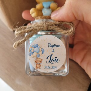 May include: A small glass jar with a cork lid and a brown twine bow. The jar is filled with small candies and has a round label with a blue and white illustration of a teddy bear holding balloons. The label says "Bapt&ecirc;me de Lo&iuml;c 29.06.2024".