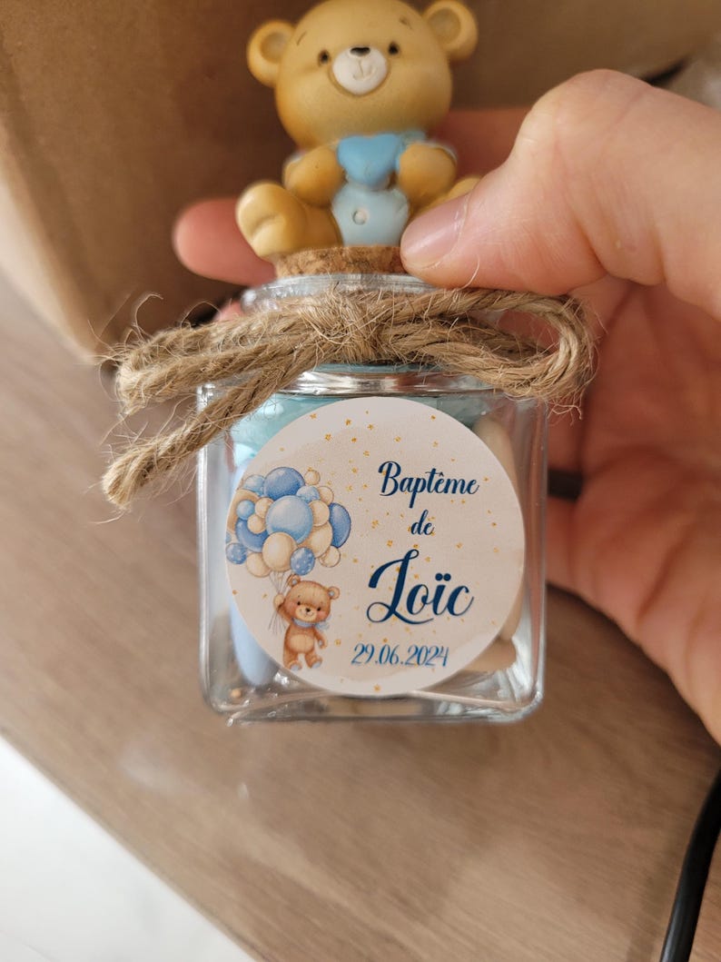May include: A small glass jar with a cork lid and a brown twine bow. The jar is filled with small candies and has a round label with a blue and white illustration of a teddy bear holding balloons. The label says "Bapt&ecirc;me de Lo&iuml;c 29.06.2024".