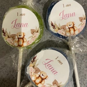 May include: Three round lollipops with a white circle sticker on each. The stickers have a brown teddy bear, pink flowers, and the text "1 an Lana".