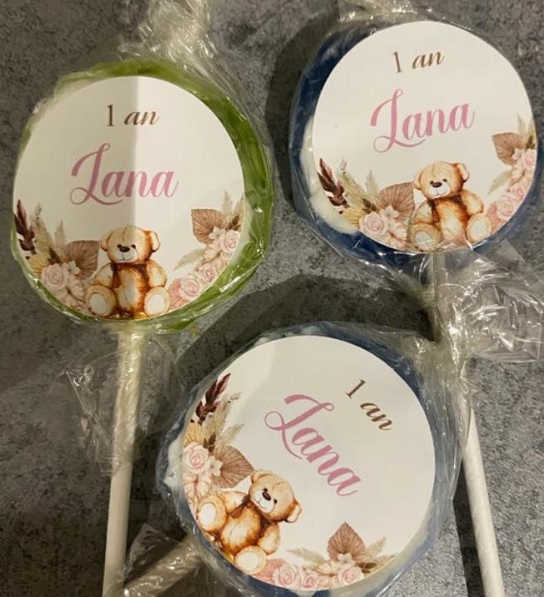 May include: Three round lollipops with a white circle sticker on each. The stickers have a brown teddy bear, pink flowers, and the text "1 an Lana".