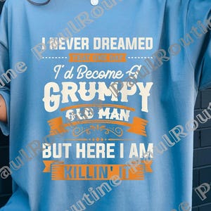 May include: Light blue t-shirt with the text "I Never Dreamed I'd Become a Grumpy Old Man But Here I Am Killin' It" in white and orange. The shirt is a relaxed fit and has short sleeves.