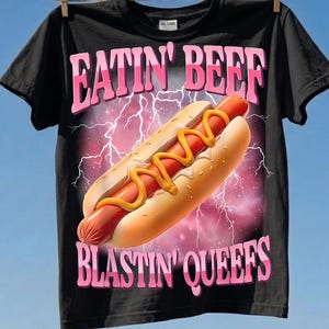 May include: A black short-sleeve t-shirt featuring a graphic of a hot dog with yellow mustard against a purple lightning background. The text EATIN' BEEF is printed in pink above the hot dog, and BLASTIN' QUEEFS is printed in pink below it.